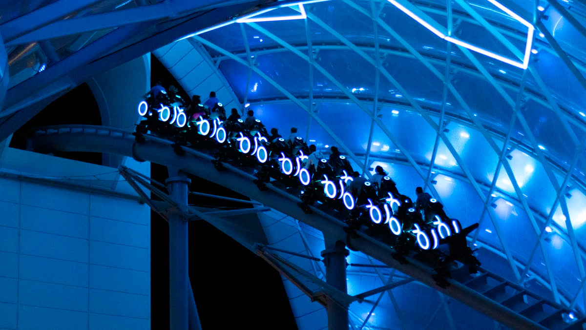 A TRON Lightcycle / Run train climbing the lift beneath the ride's blue-lit canopy at night, riders leaning forward on the lightcycles